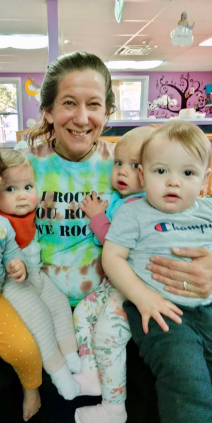Smiling daycare teacher holds three toddlers indoors.