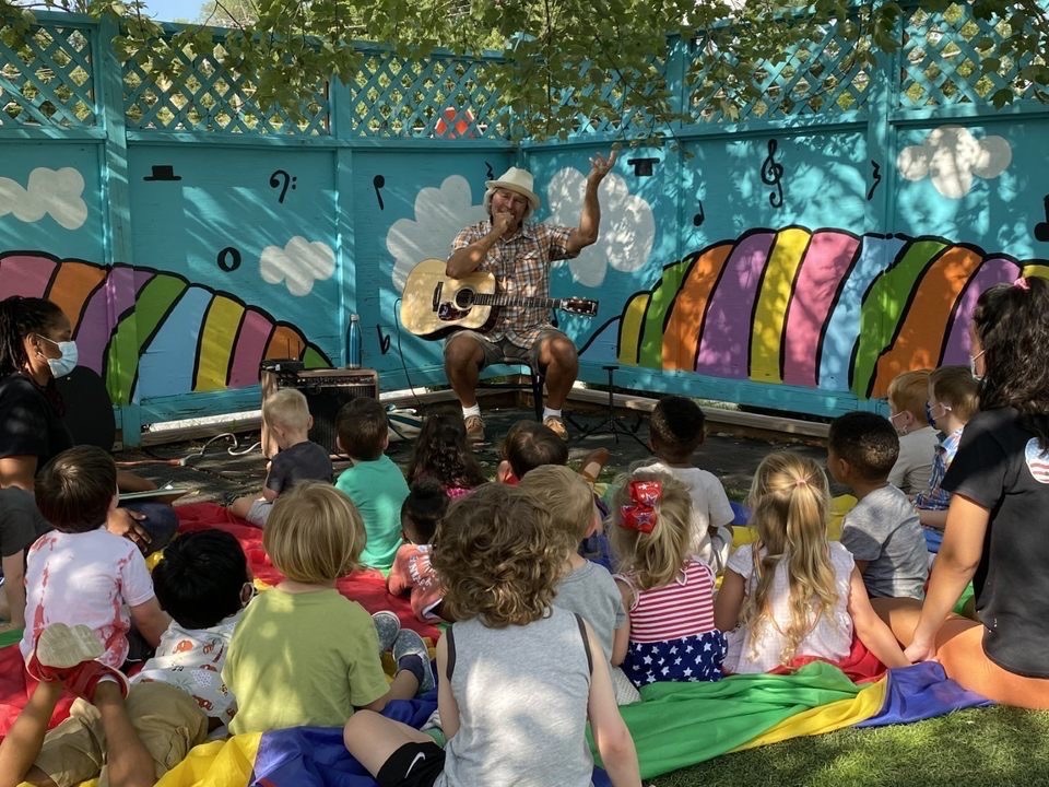 Guitarist performs for children in colorful outdoor setting.