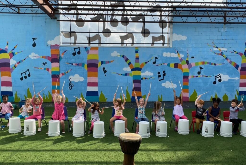 Children drumming on buckets against colorful mural backdrop.