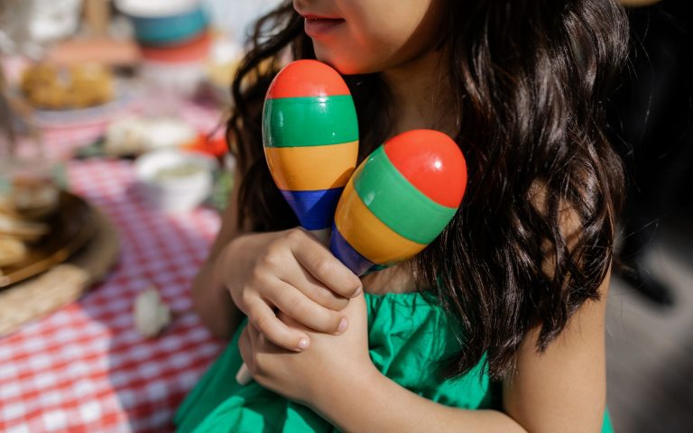 A playful child from a music-integrated preschool, in a dress holds colorful maracas
